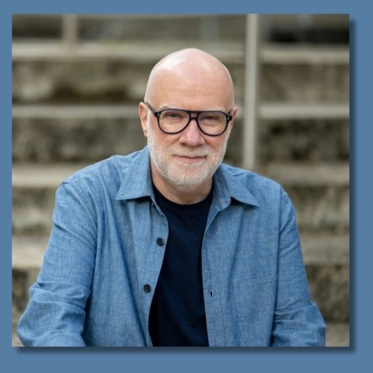 A colour portrait of John Spencer Williams, a white man with grey beard and glasses, who is wearing a blue shirt.